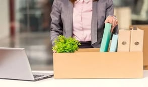 A woman is packing away her belongings from work into a box at her desk. In the box, she has a plant and several folders.